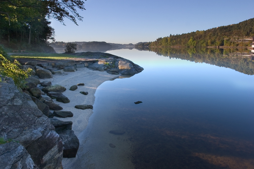 Fotografi av en innsjø med strand og svaberg i forgrunn. Innsjøen er ellers omkranset av trær som ligger i et kuppert landskap.